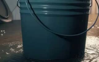 A bucket on a wet floor with water leaking indoors, highlighting the issue of basement drain backup and the need for prompt cleanup and repair.