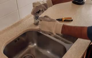 A plumber repairing a kitchen sink faucet with gloves, emphasizing the benefits of water leak detectors in preventing water damage and ensuring early detection.