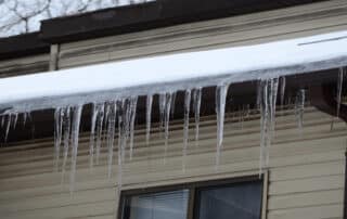 Snow-covered roof with long icicles hanging from the gutter above a window on a beige house, indicating potential ice dam formation.