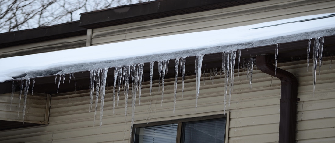 Snow-covered roof with long icicles hanging from the gutter above a window on a beige house, indicating potential ice dam formation.