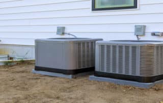Two outdoor HVAC units installed on concrete pads next to a white house, used for air conditioning and cooling systems.