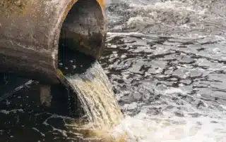Dirty water flowing from a pipe into a river near sewage treatment facilities, showing environmental pollution after sewage mitigation. Image represents lingering odor sources such as contaminated water, organic waste, and runoff that continue to produce unpleasant smells even after cleanup.