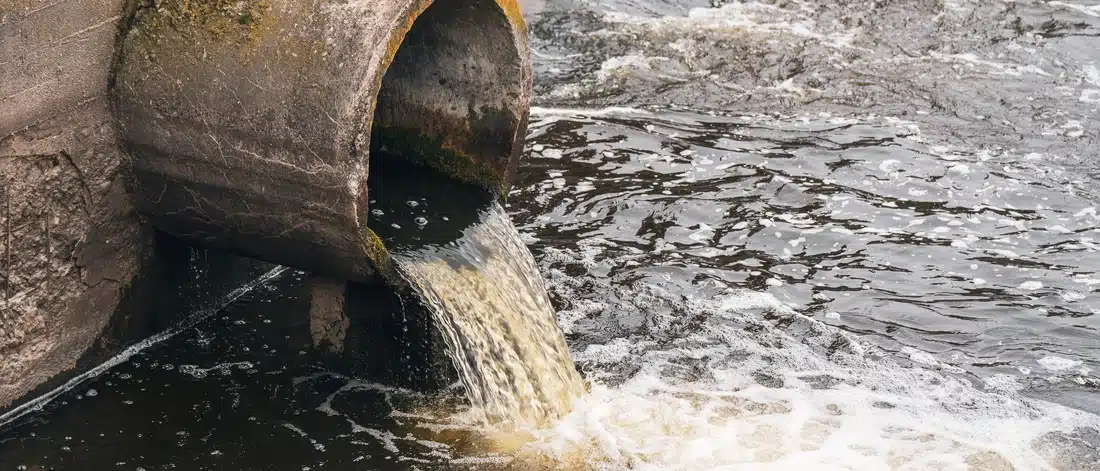 Dirty water flowing from a pipe into a river near sewage treatment facilities, showing environmental pollution after sewage mitigation. Image represents lingering odor sources such as contaminated water, organic waste, and runoff that continue to produce unpleasant smells even after cleanup.