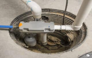 An overhead, close-up shot of a sump pump system installed in a circular basin within a concrete floor. The image shows PVC piping, a blue flexible hose, and a gray control unit with a float switch submerged in a small amount of water.