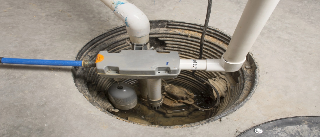 An overhead, close-up shot of a sump pump system installed in a circular basin within a concrete floor. The image shows PVC piping, a blue flexible hose, and a gray control unit with a float switch submerged in a small amount of water.