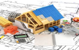 Model of a house under construction placed on architectural blueprints, surrounded by building materials, tools, paint cans, calculator, and notebook representing restoration planning and rising material costs.